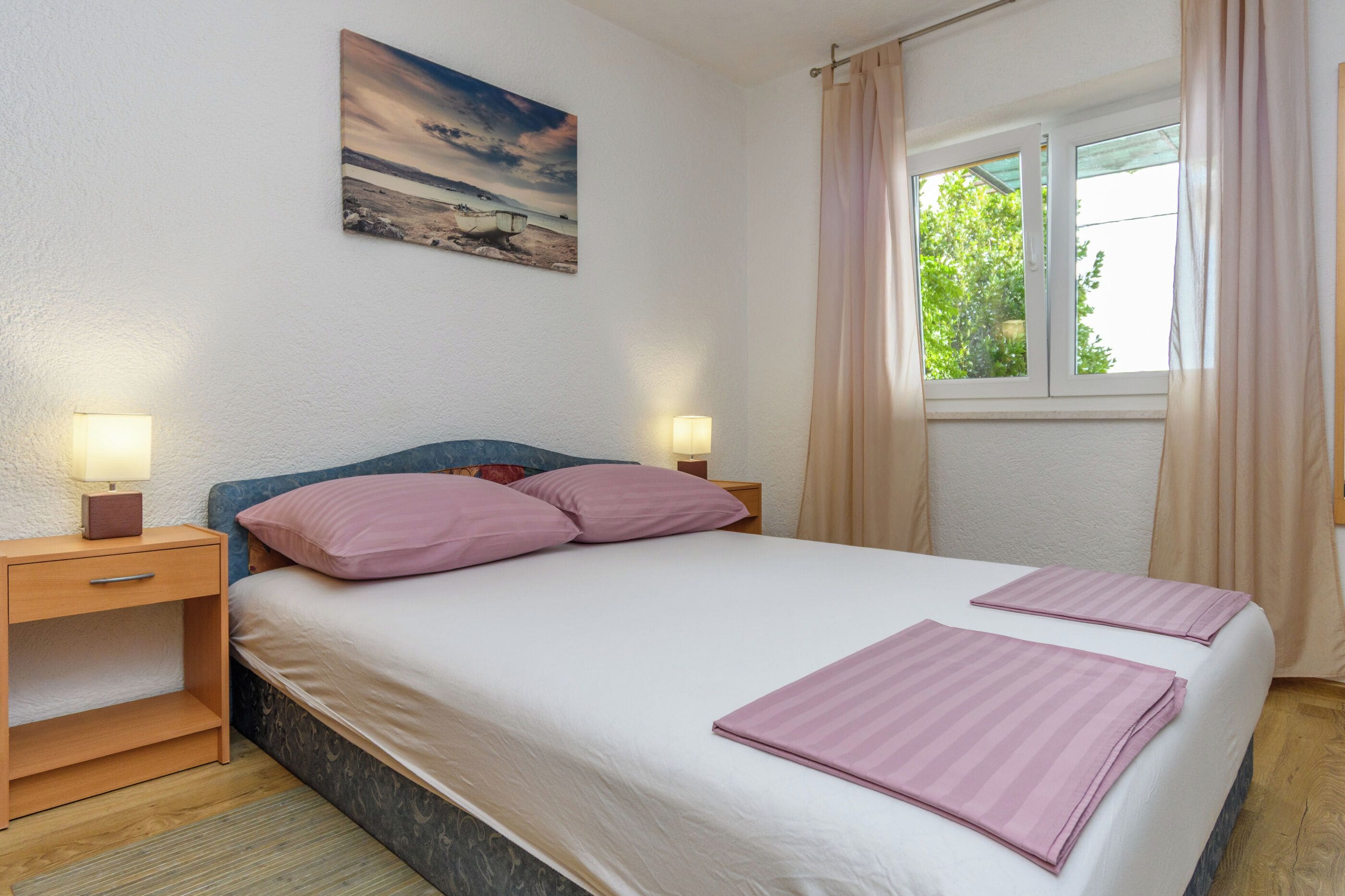 A bedroom with a bed, two bedside tables, a nightstand, a window with curtains, and a painting on the wall.