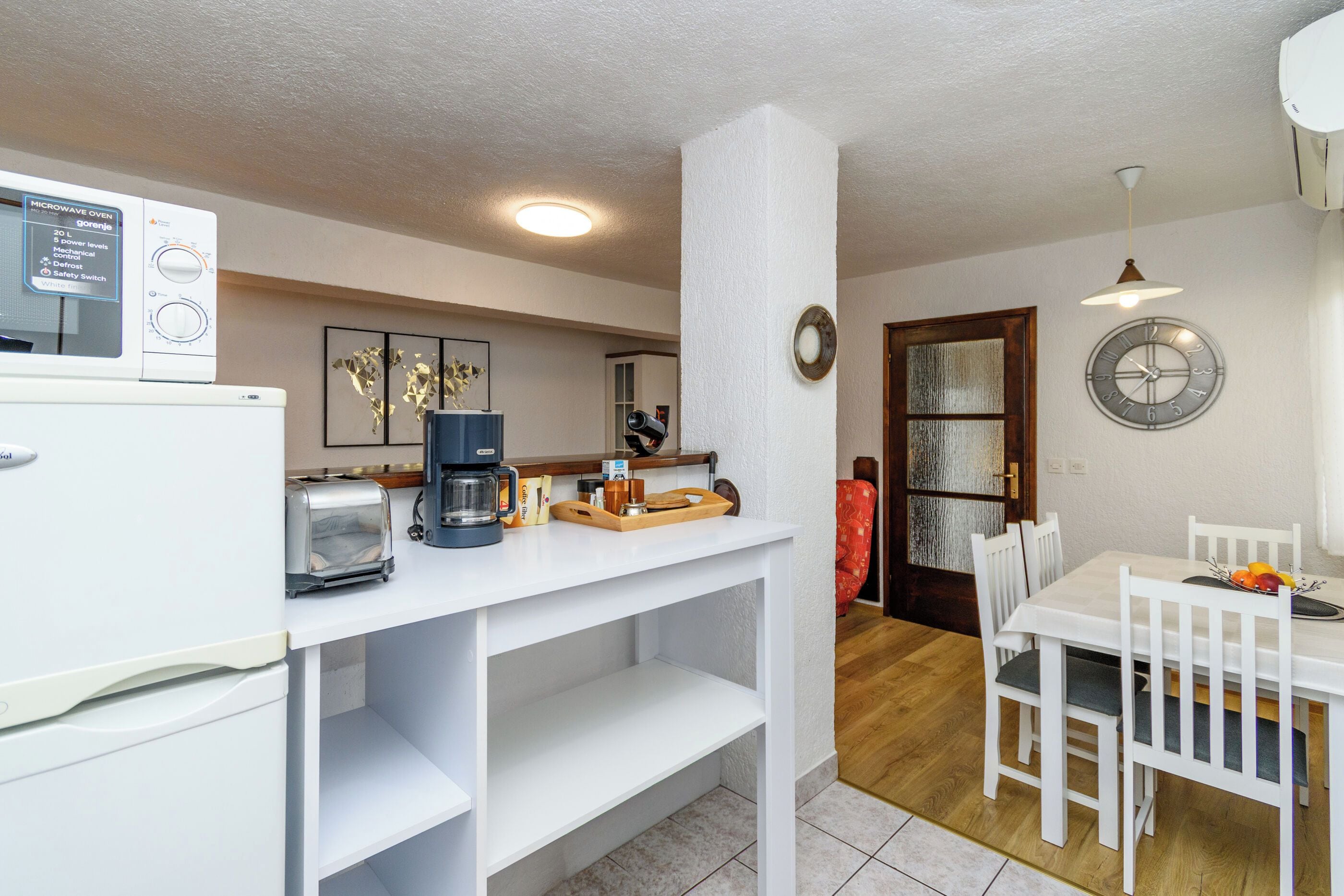 Een kleine keuken met een magnetron, broodrooster en koffiezetapparaat op het aanrecht, een eethoek met een tafel en stoelen, en een klok aan de muur.