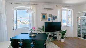 A modern living room with a dining area, large windows with a view of the sea, a flat-screen TV, and a glass cabinet with decorative items. - Apartment Ljerka (Postira)