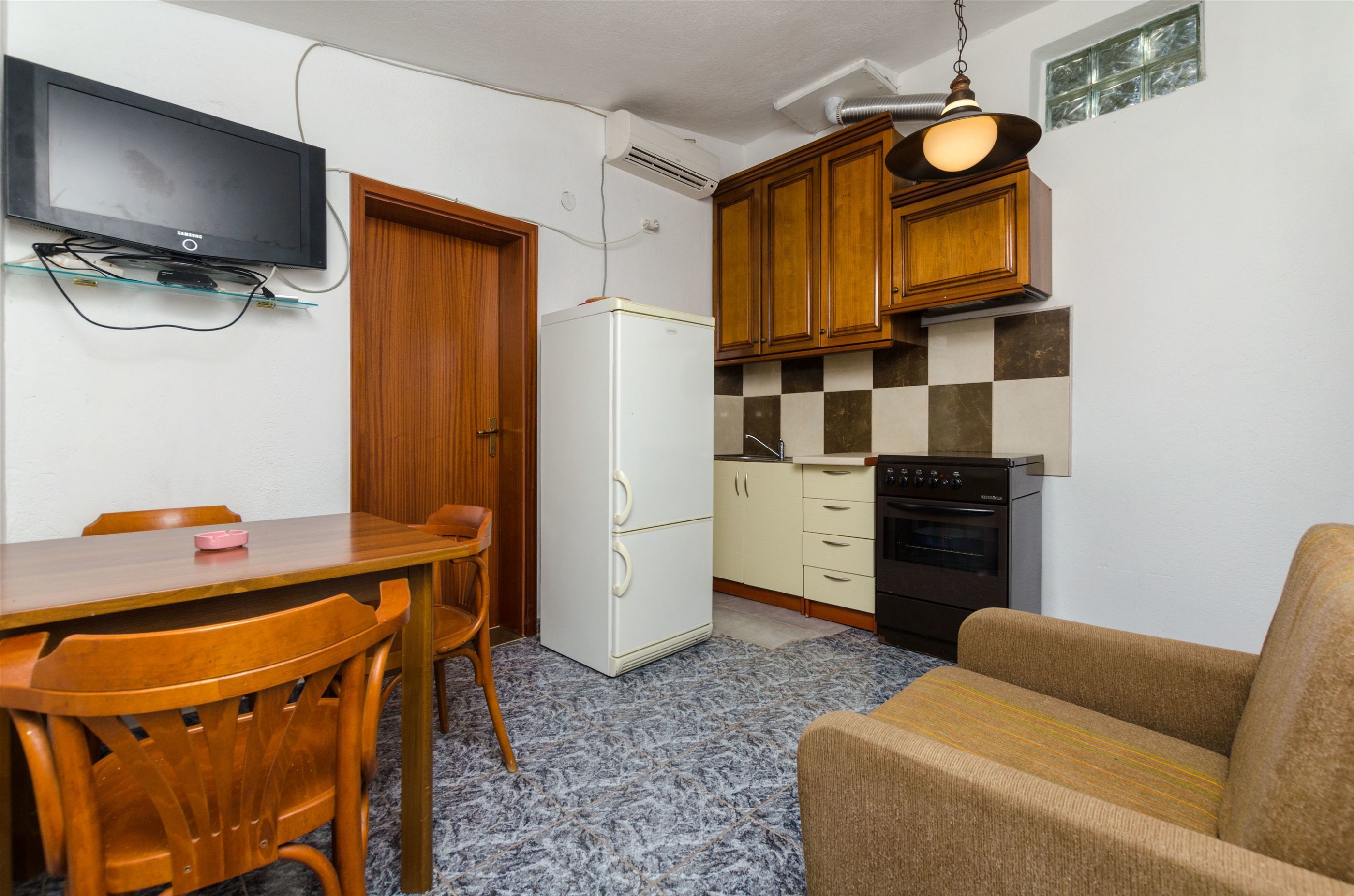 Een kleine keuken met een koelkast, oven en houten keukenkastjes. Er is een eettafel met stoelen en een televisie aan de muur bevestigd.