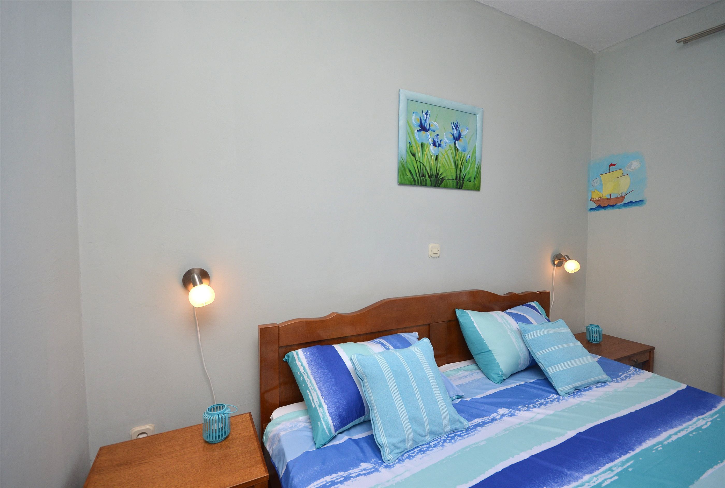 Een slaapkamer met een houten hoofdbord, een bed met blauw-witte strepen lakens, twee wandlampen voor het lezen en een klein tafeltje met een blauwe vaas.