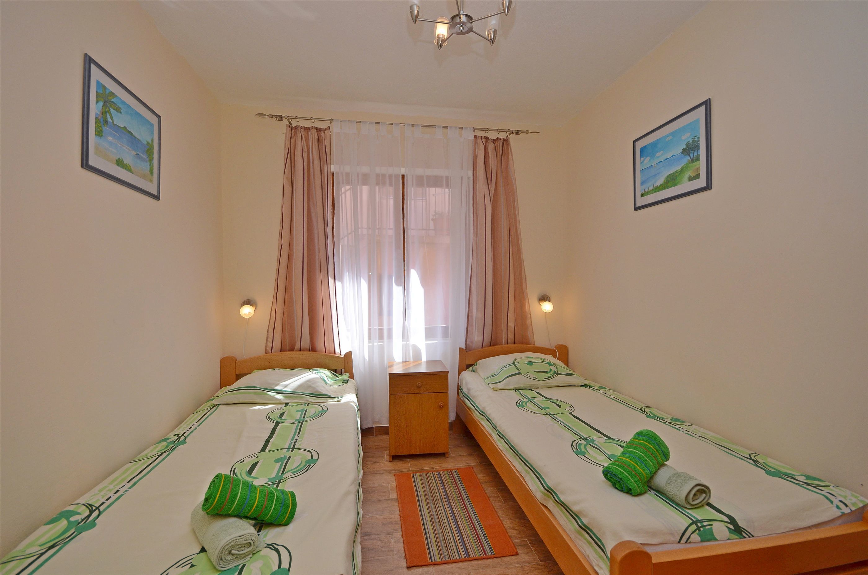 A room with two single beds, a nightstand, a small table, and a window with sheer curtains.