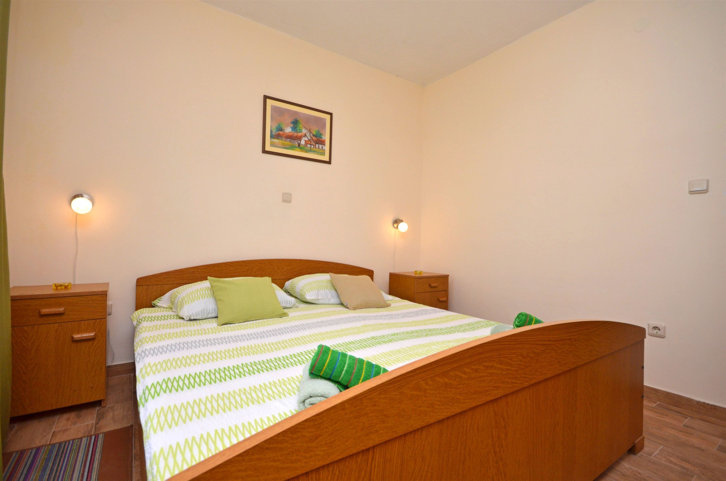 A bedroom with a wooden bed, two bedside tables, a nightstand, a painting on the wall, and two wall-mounted lights.