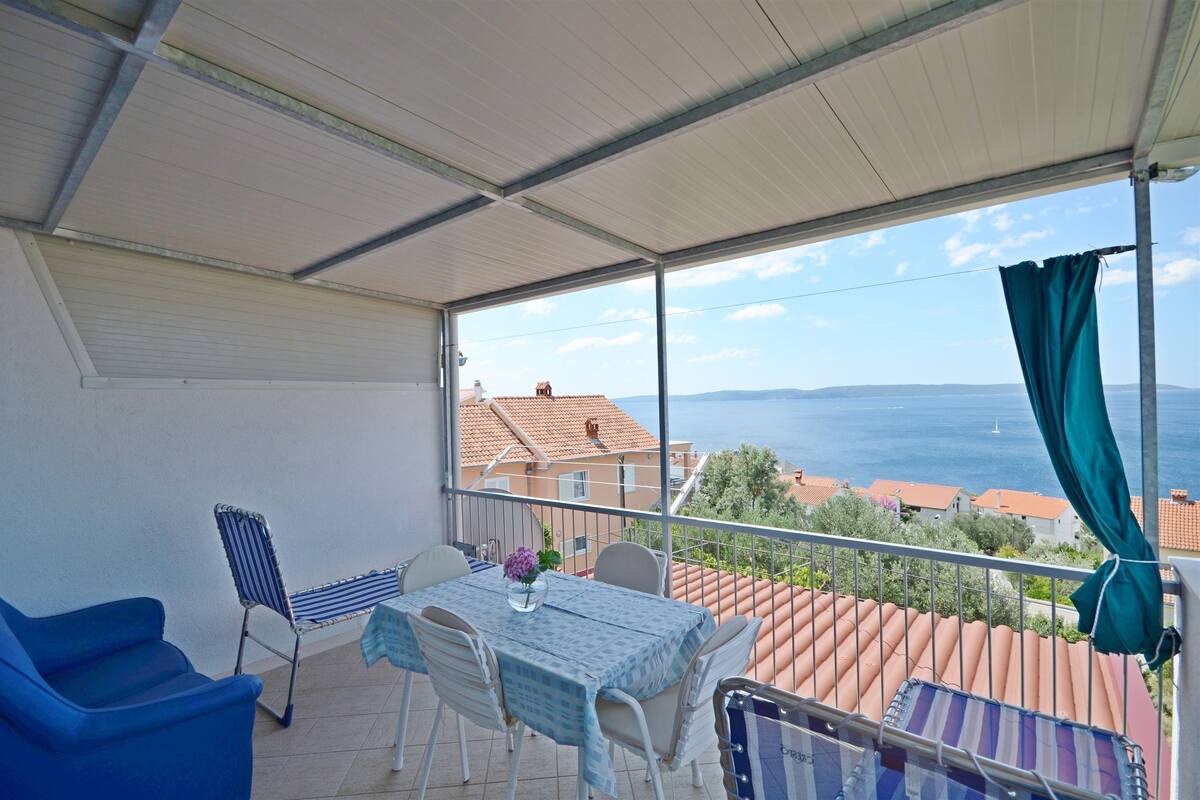 A balcony with a table and chairs, a view of the sea, and a building with red-tiled roofs in the background.