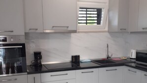 A modern kitchen with white cabinets, a built-in oven, and a window with blinds. - Apartments Grozdana (Postira)