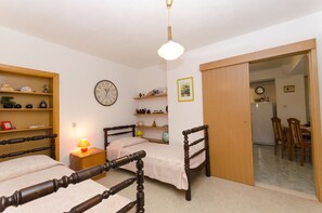 A bedroom with two wooden beds, a bedside table with a lamp, a clock on the wall, and a door leading to a dining area.