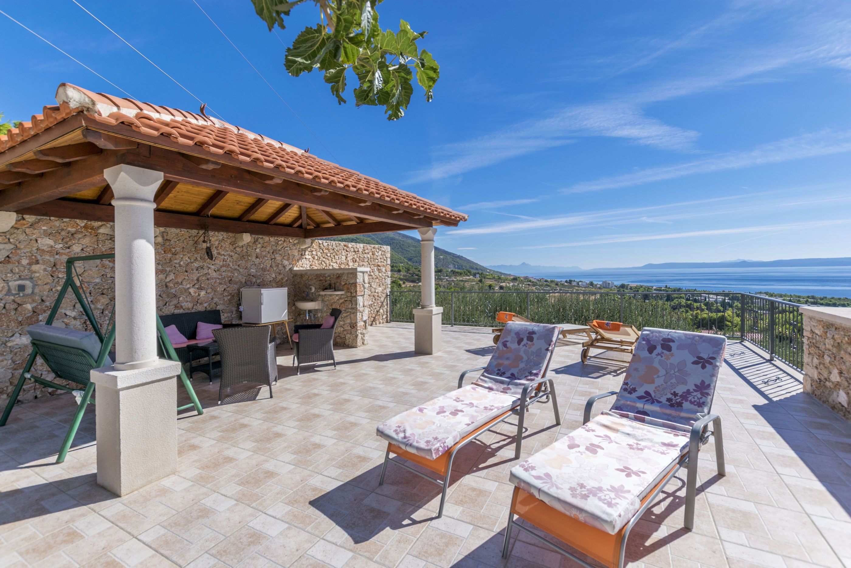 Une terrasse avec un mur en pierre, une pergola, des chaises longues et une vue sur la mer.