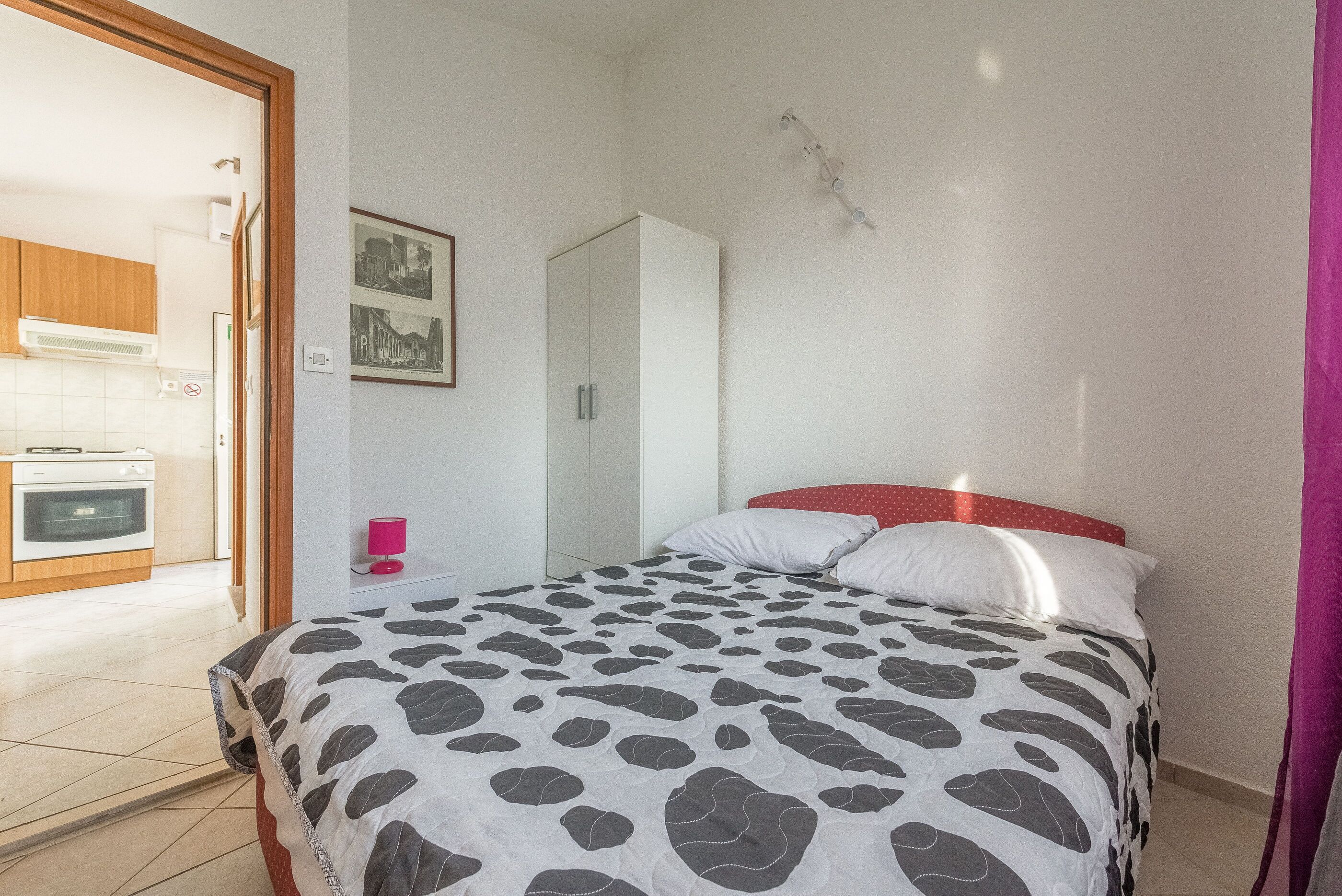A bedroom with a bed, a wardrobe, a mirror, and a door leading to a kitchen.