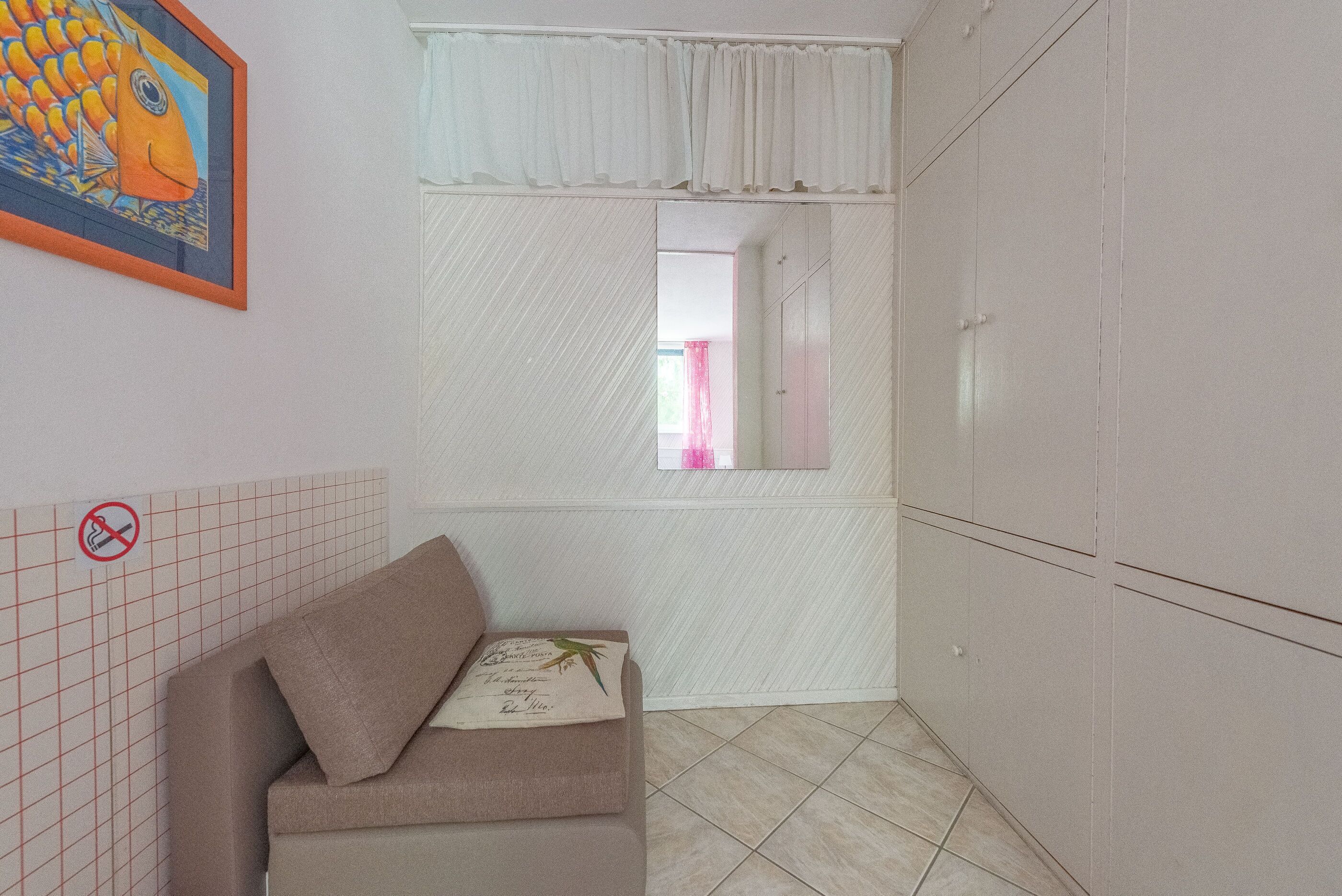 A small, private bathroom with a white tiled floor, a beige sofa with a pillow, a framed fish painting, and a mirror above the sink.
