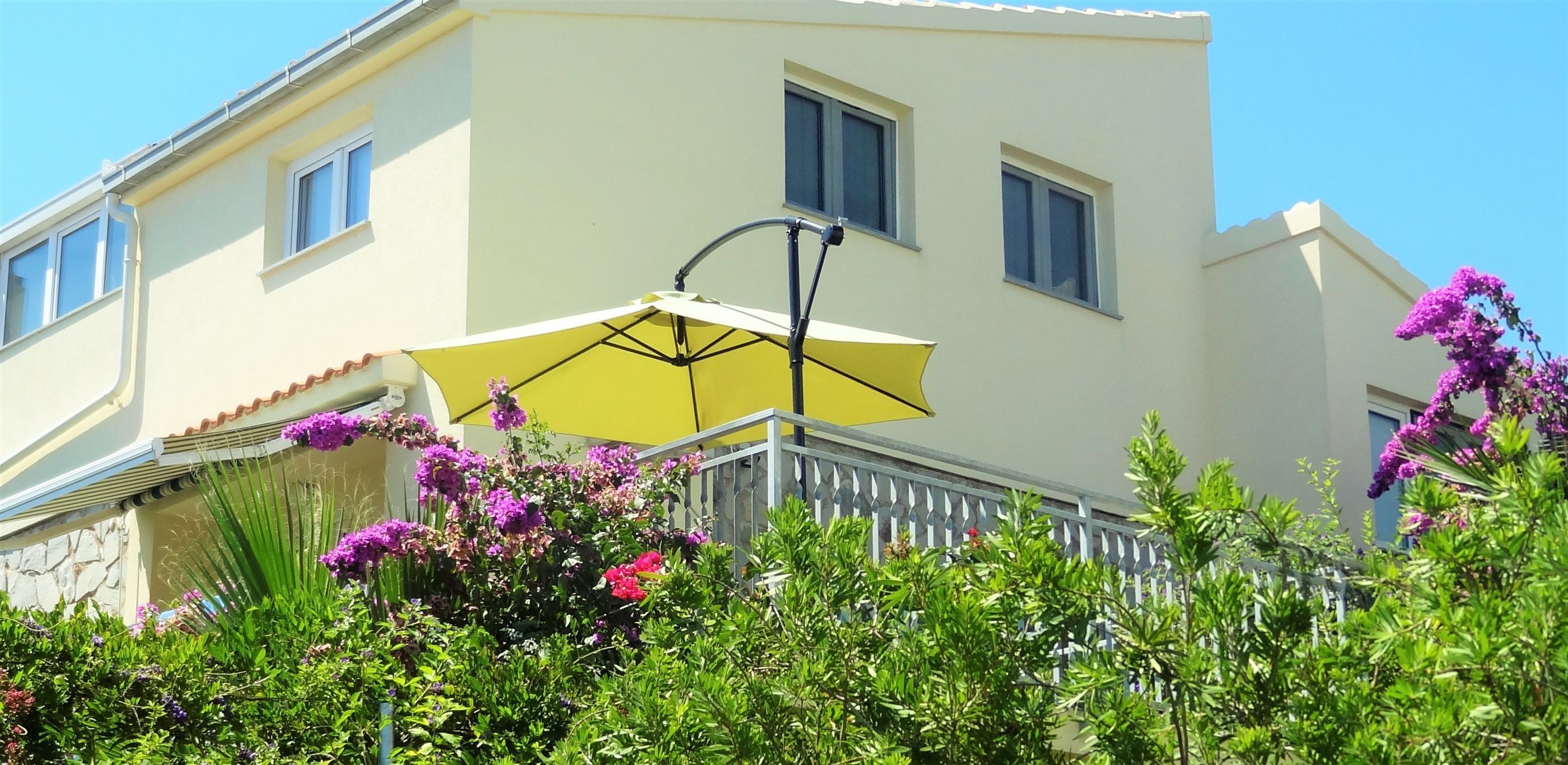 Ein zweistöckiges Gebäude mit Balkon, gelber Sonnenschirm und violetten Blumen.