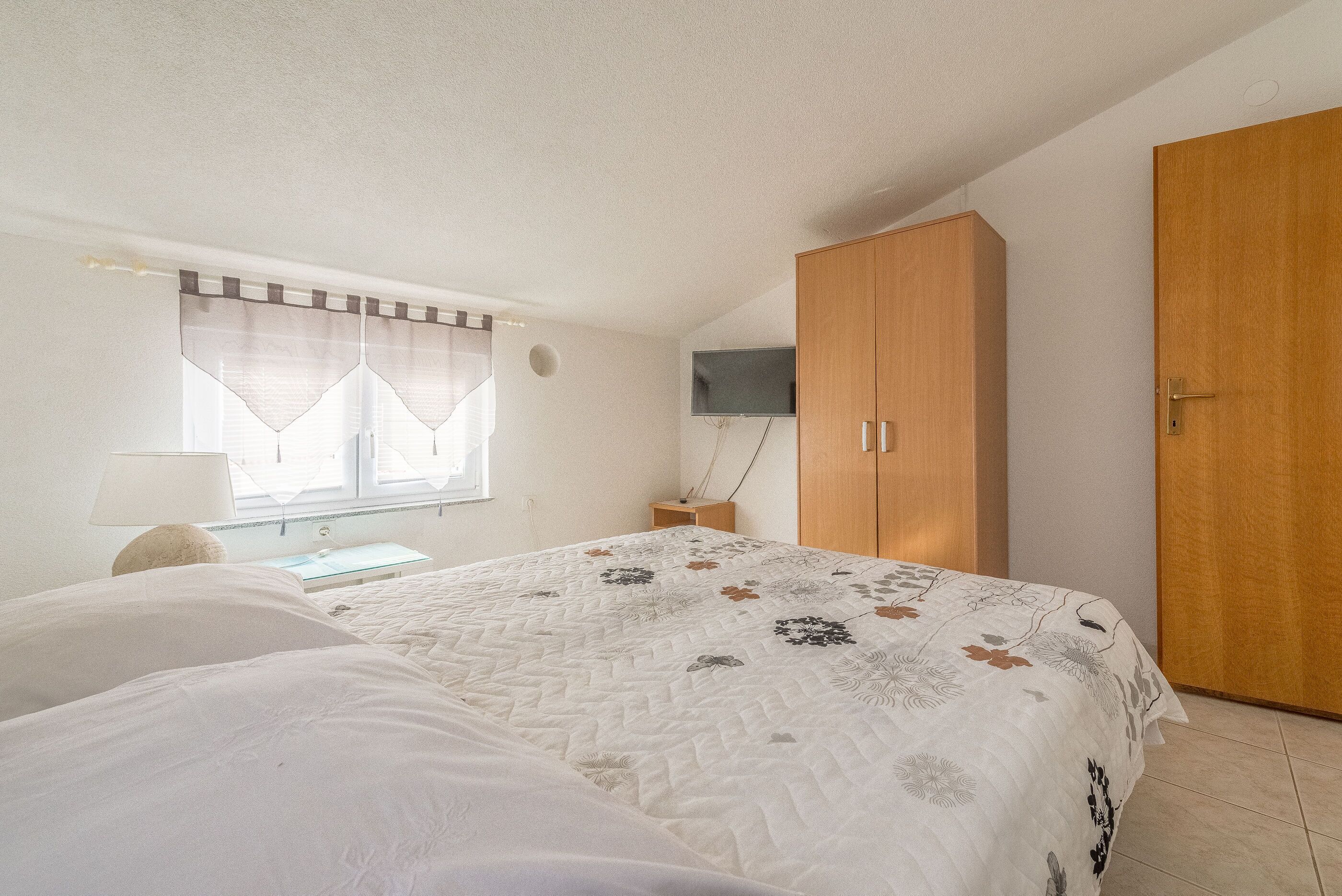 Ein Schlafzimmer mit einem Bett, einem Holzschrank, einem an der Wand montierten Fernseher und einem Fenster mit Vorhängen.