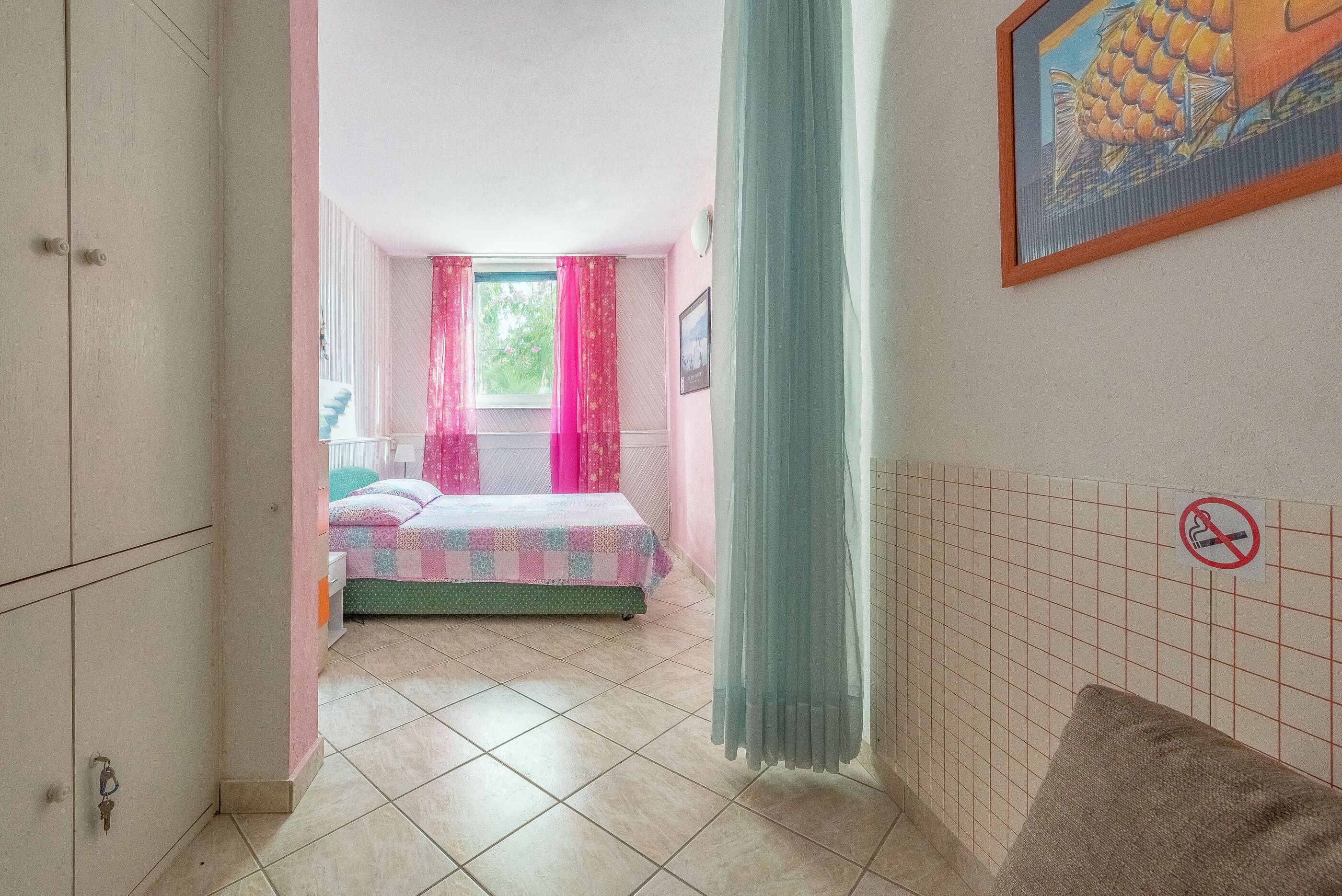 Una camera da letto con un letto, una finestra con tende rosa, un quadro al muro e un cartello ‘divieto di fumo’.