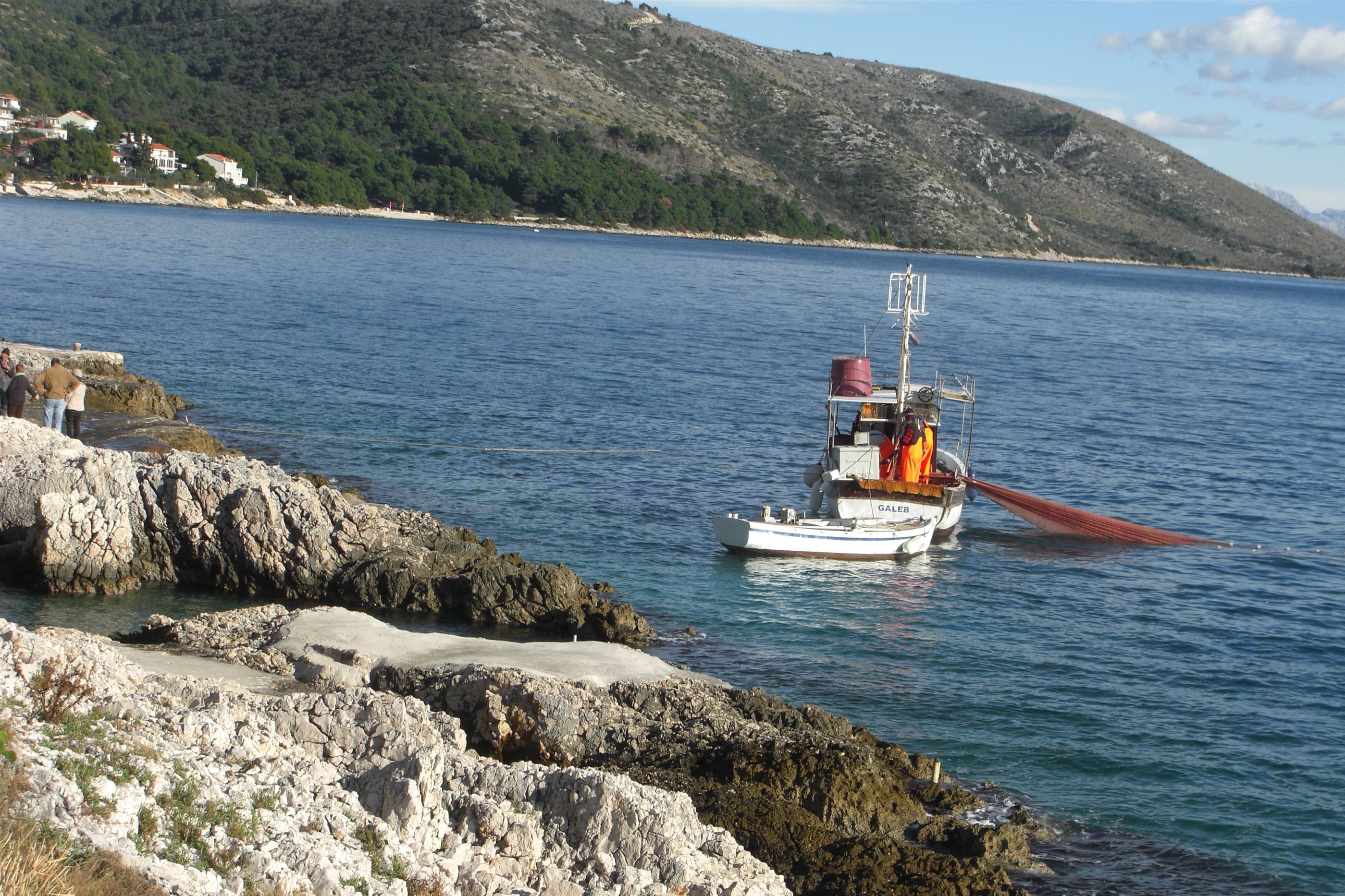 Ein Boot namens GALLEB liegt vor einer Küste mit felsigem Ufer und einem Bergpanorama vor Anker.