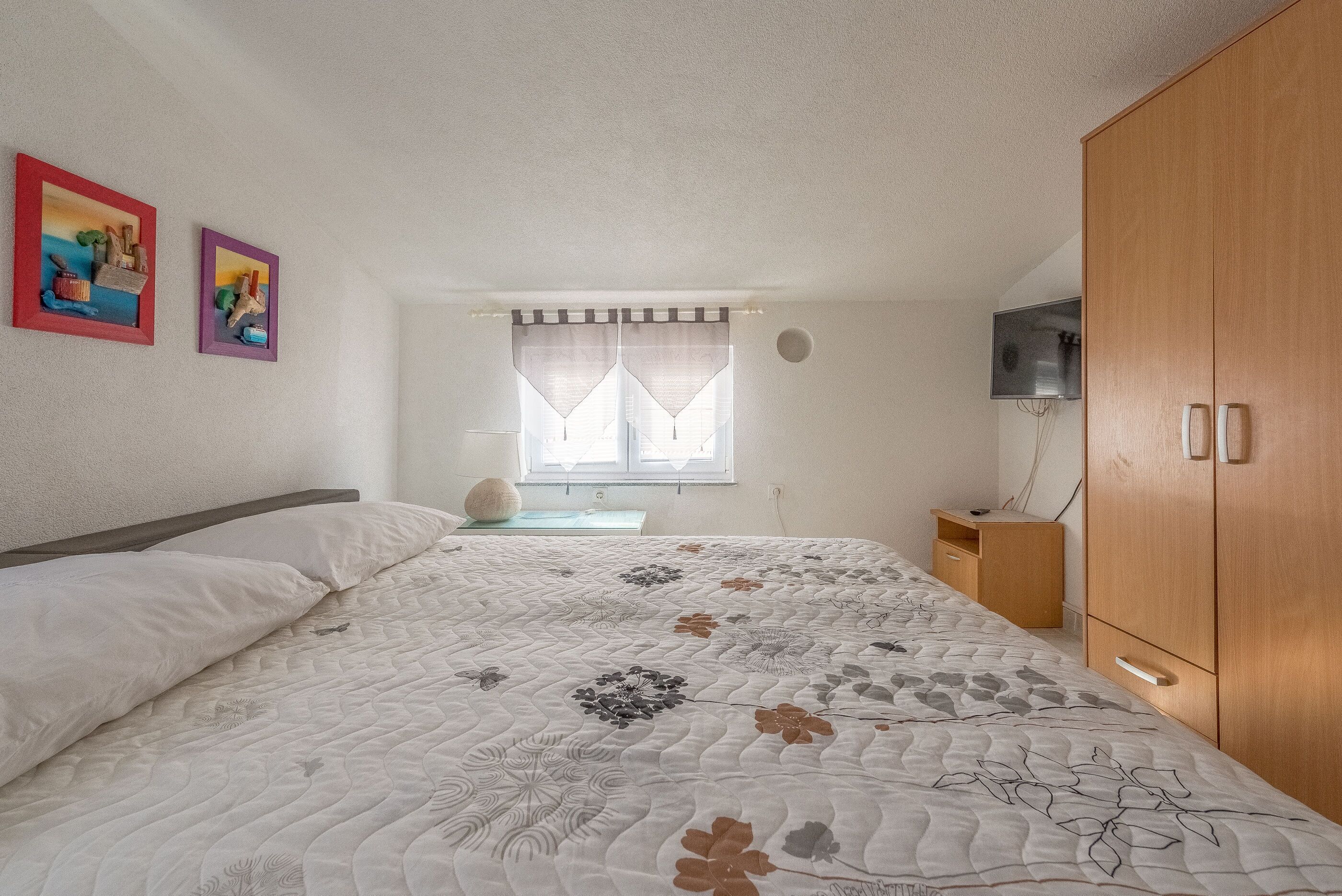 A bedroom with a bed, a wooden wardrobe, a TV on the wall, and two framed pictures on the wall.