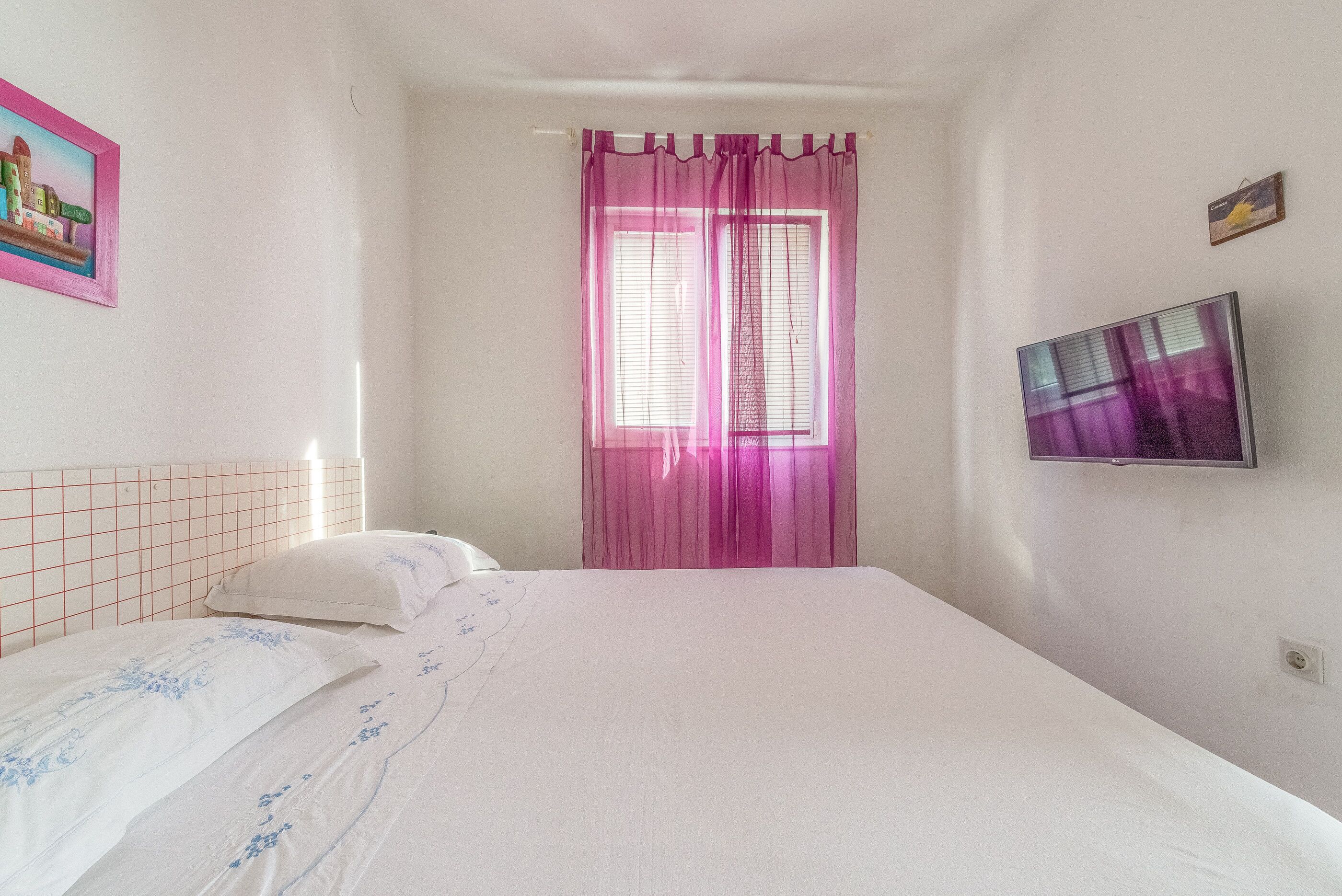 Un dormitorio con cama, ventana con cortinas rosadas, televisor en la pared y un pequeño marco de fotos.