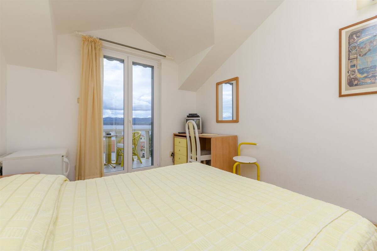 A bedroom with a bed, a desk, a chair, a radiator, a window with curtains, and a framed picture on the wall.