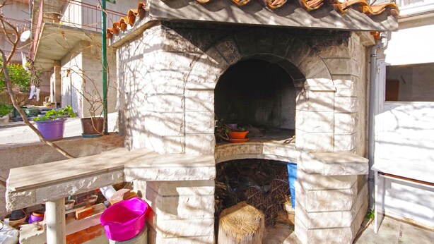 A stone-built outdoor oven with a tiled roof, surrounded by a concrete base and a wooden bench. - Apartment Danica (Makarska)
