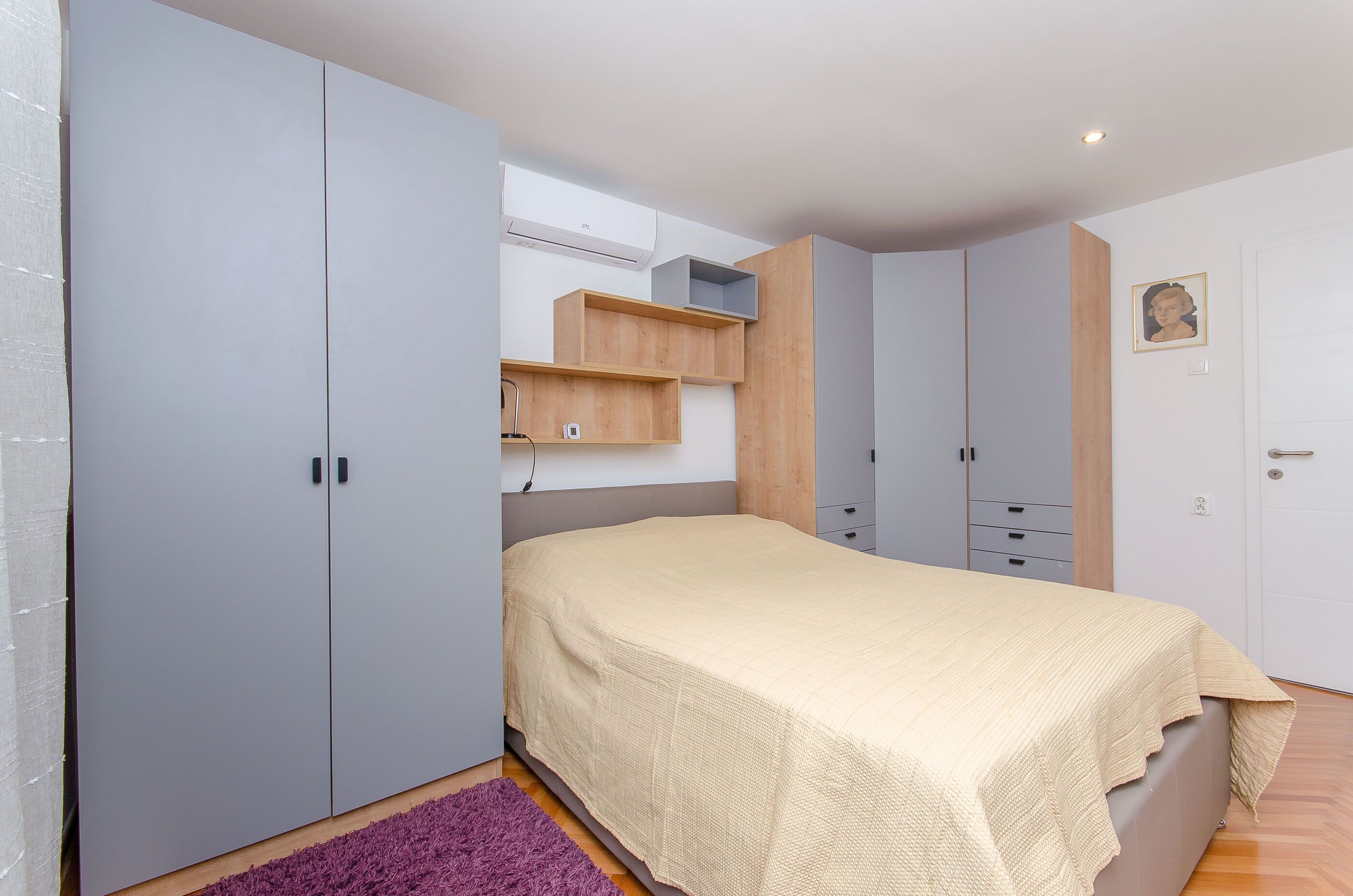 A bedroom with a bed, grey wardrobe, wooden shelves, and a purple rug.