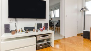 A modern living room with a large flat-screen TV mounted on the wall, a white entertainment unit with speakers and a small shelf, and a wooden floor. - Apartment Sunny (Split)
