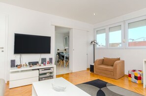 Una sala moderna con un televisor de pantalla plana, un sofá, una mesa de centro y un comedor visible a través de un puerta abierta.