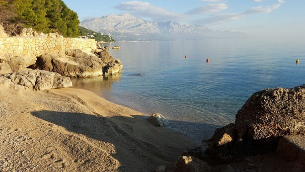 A rocky beach with clear water, a stone wall, and mountains in the background. - House Ivan (Mimice)