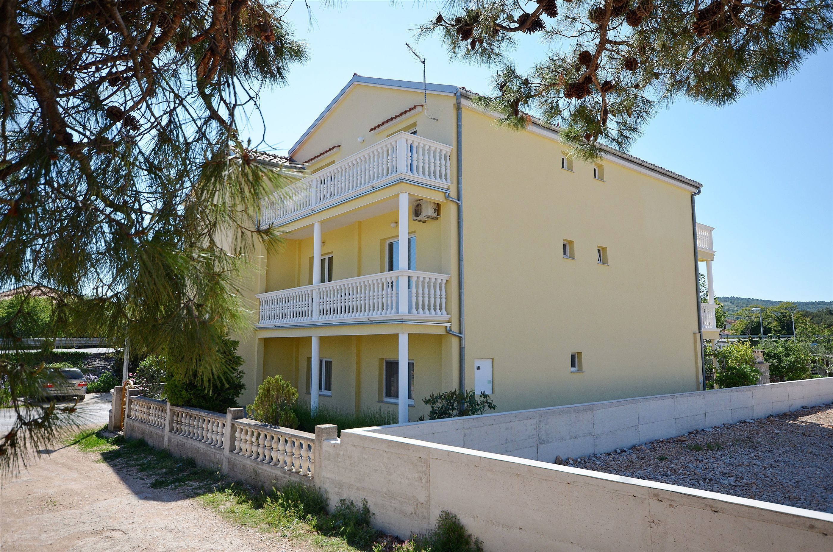 A two-story building with a balcony on the upper floor, surrounded by trees and a gravel path.
