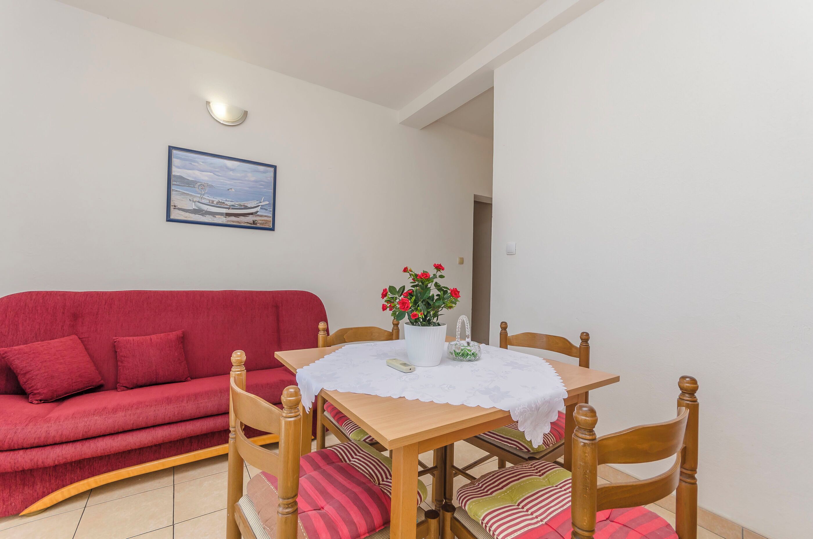 Una sala acogedora con un sofá rojo, una mesa de comedor de madera con mantel blanco y una planta en maceta con flores rojas.