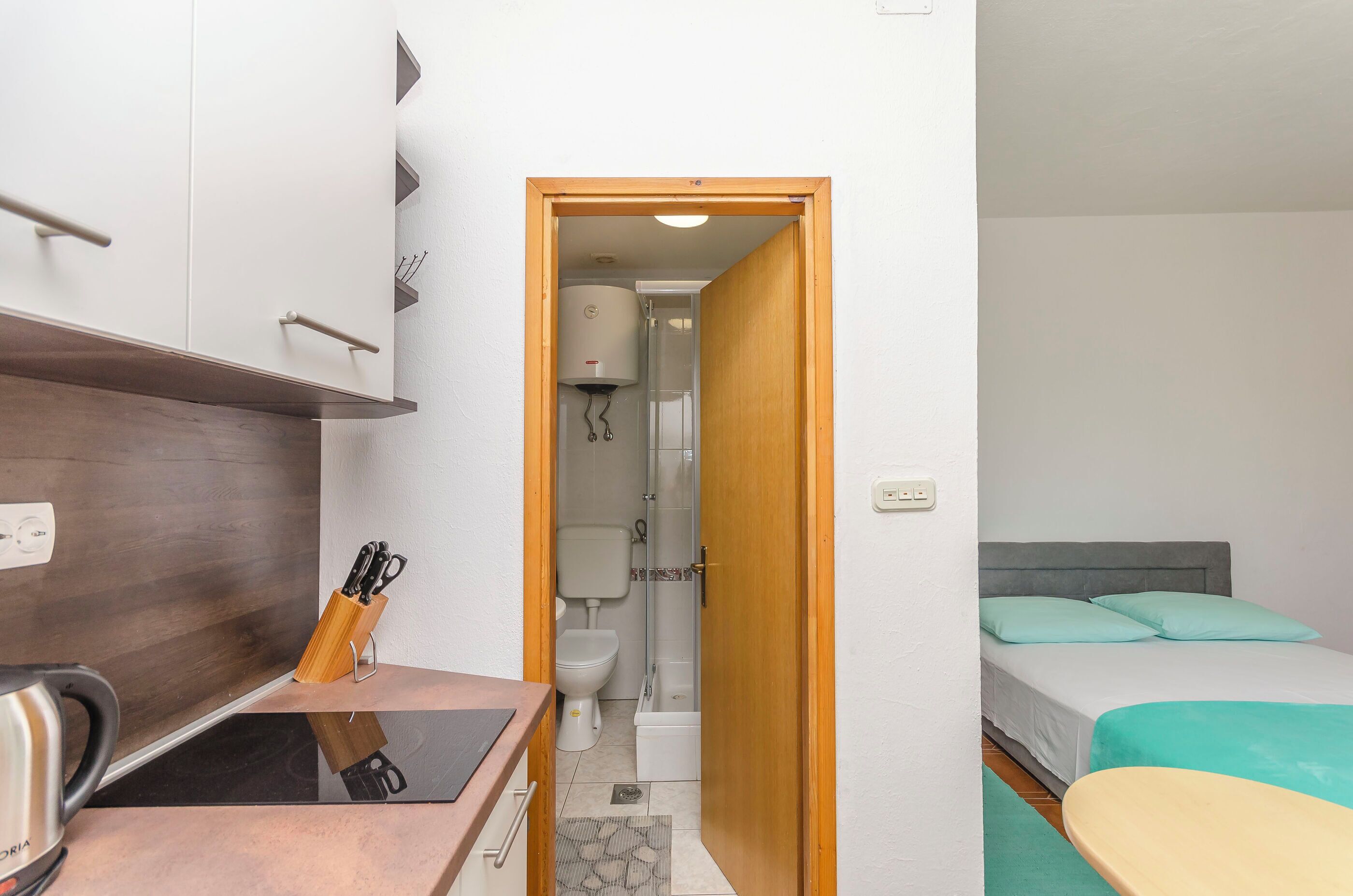 A compact hotel room with a wooden cabinet, a small kitchenette, a bed with teal bedding, and a shower area.