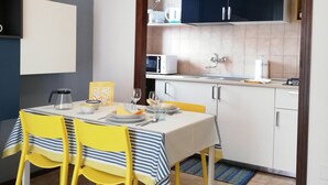 A dining area with a table set for two, yellow chairs, a white tablecloth, and a kitchen in the background with cabinets and appliances. - Apartment Ivrea center - UNESCO site (Ivrea)