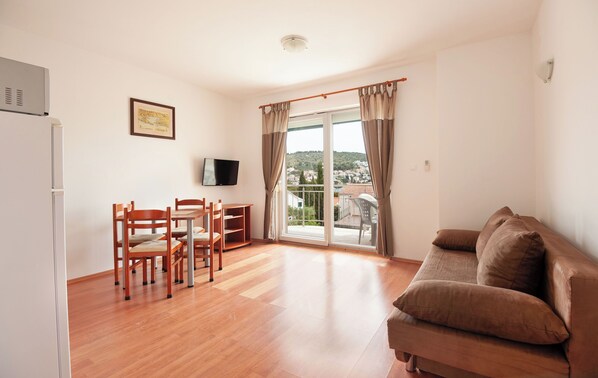 A living room with a wooden floor, a brown sofa, a dining table with chairs, a television, and a balcony with curtains.