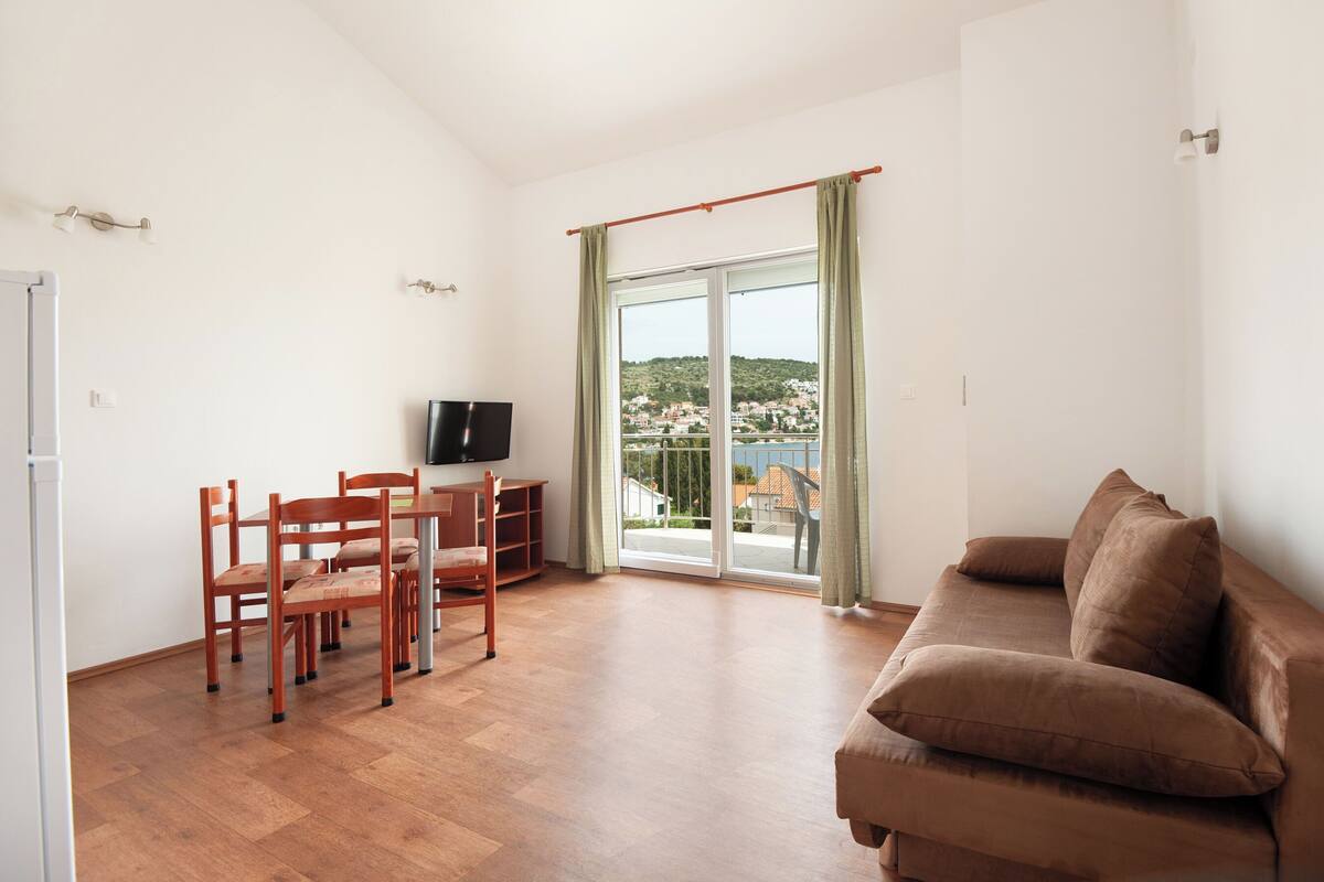 A living room with a brown sofa, wooden chairs, a TV, and a sliding glass door leading to a balcony with a view of a hillside.