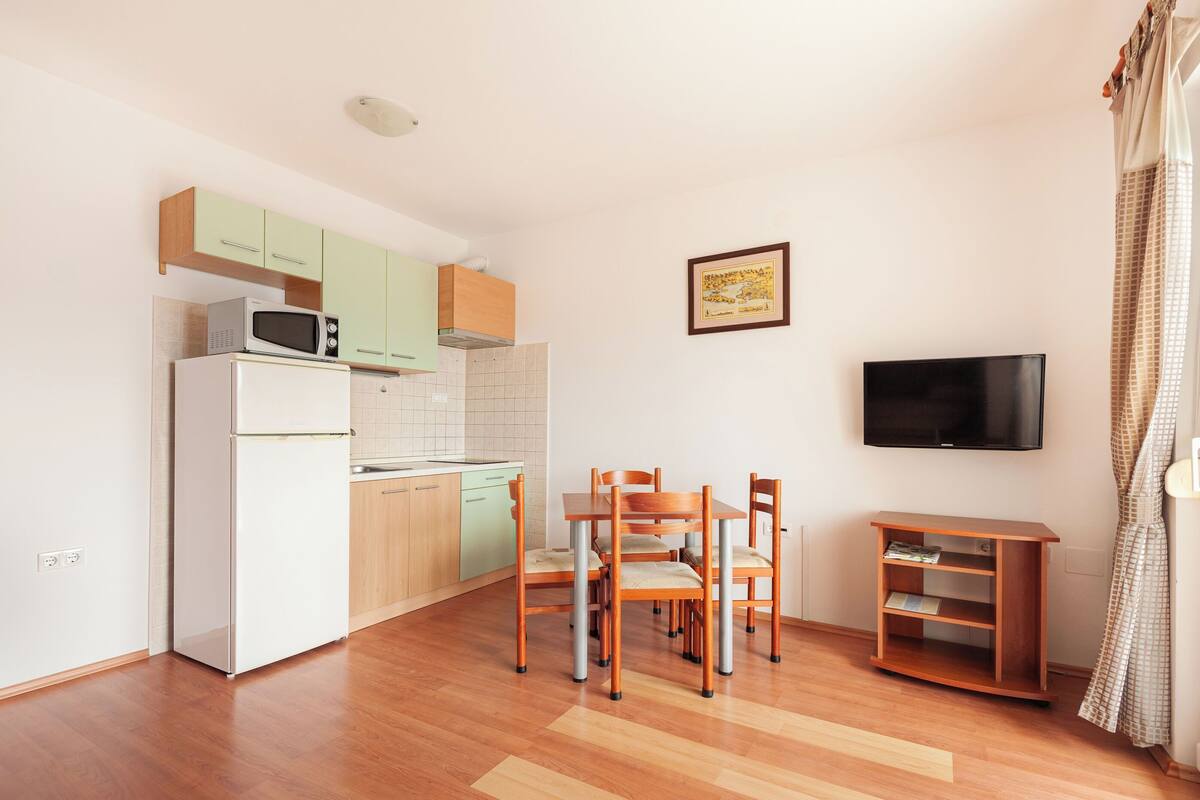 A compact kitchen with a microwave, refrigerator, and wooden cabinets. There is a dining table with chairs, a television on the wall, and a small wooden side table.