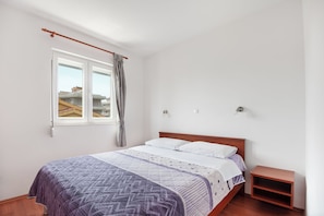 A bedroom with a bed, a window with curtains, a bedside table, and a small wooden shelf.