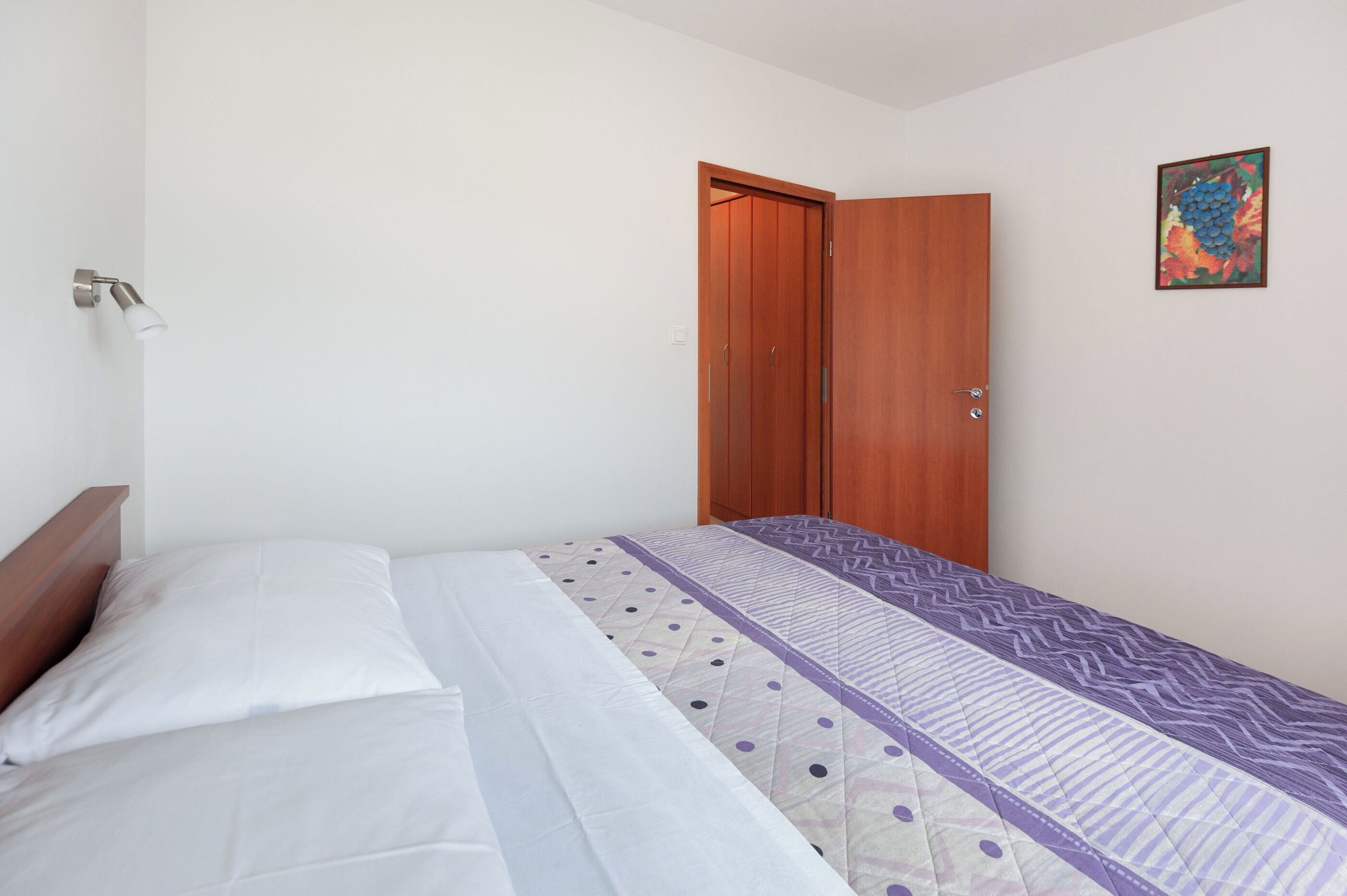 A bedroom with a bed, a wooden wardrobe, a wall-mounted light, and a framed picture.