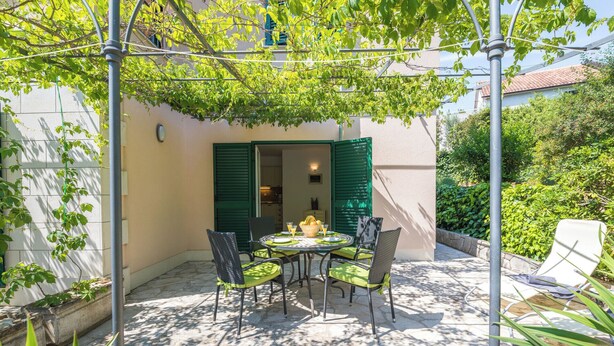 Un patio con tavolo e sedie, un pergolato con piante rampicanti e vista su un edificio con persiane verdi.