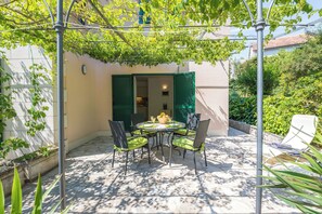 Eine Terrasse mit Tisch und Stühlen, ein mit Kletterpflanzen bewachsener Pergola und Blick auf ein Gebäude mit grünen Fensterläden.