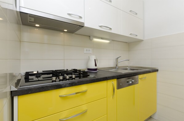 A kitchen with yellow cabinets, a black countertop, a stove, a kettle, and a sink. - Apartments Karmela (Primosten)