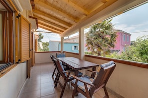 En overdækket balkon med et træbord og stole, udsigt til en lyserød bygning og en palme.