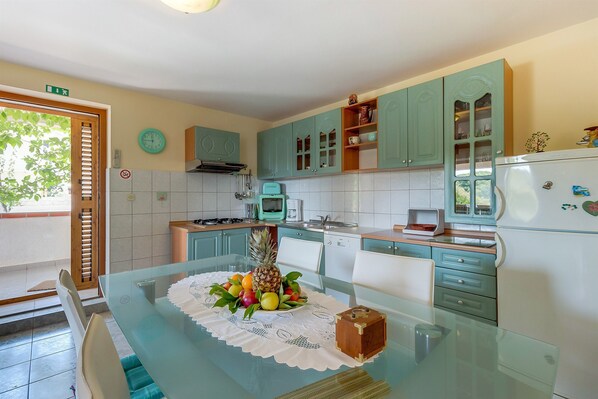 Une cuisine avec des meubles de couleur turquoise, une table en verre agrémentée d’un centre de table composé de fruits et une horloge au mur.