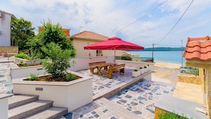 En terrasse med et træbord og rød parasol, trapper der fører op til en balkon med udsigt over en strand.
