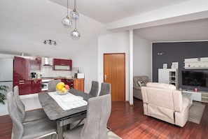 A modern kitchen with red cabinets, a dining table with chairs, a sofa, and a television. - Apartments Zivana (Primosten)