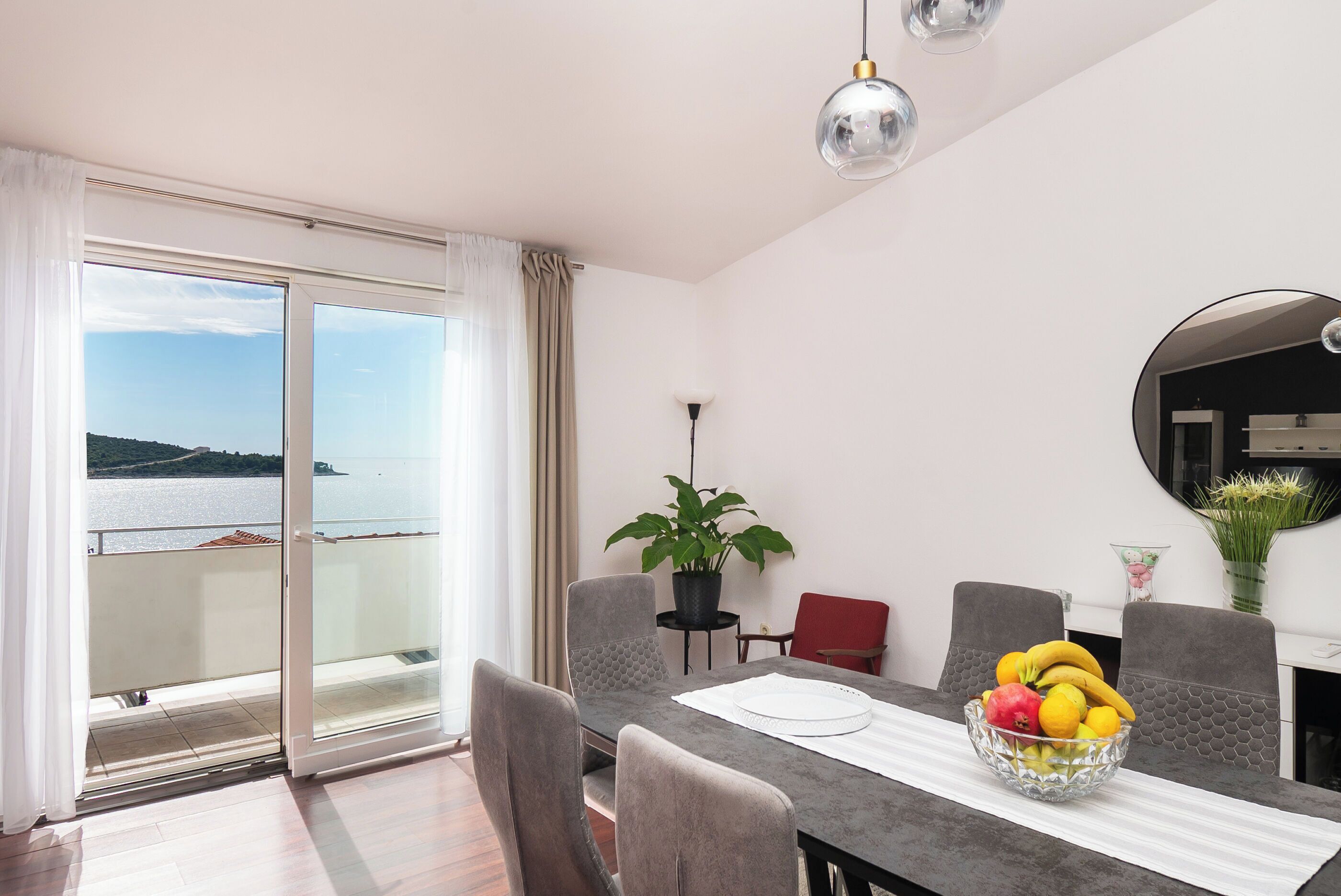 A dining area with a table, chairs, a bowl of fruit, a potted plant, and a view of the sea through a sliding door.