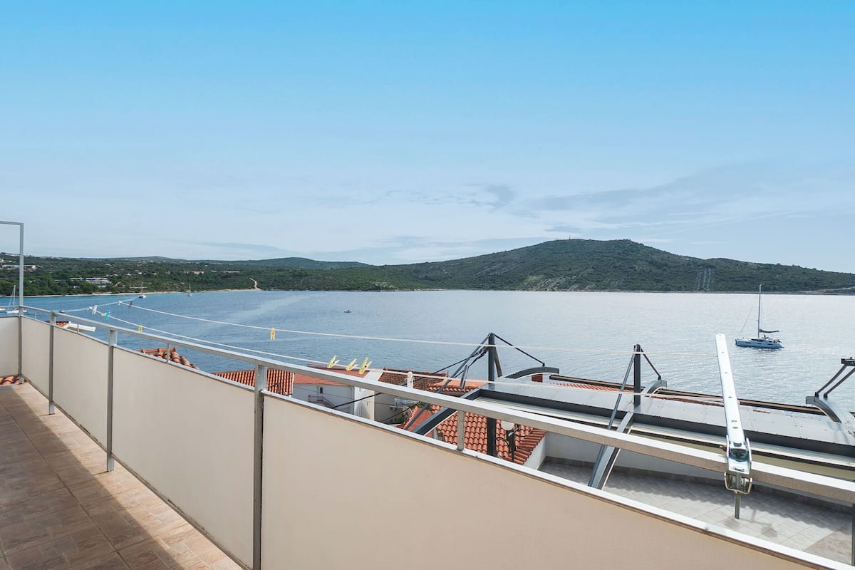 A balcony with a view of a calm body of water, a sailboat, and distant hills.