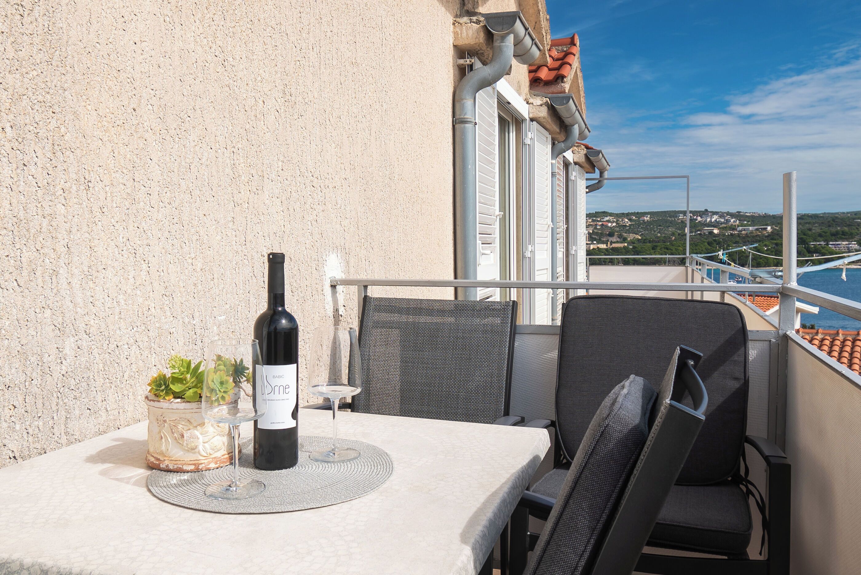 A balcony with a table set for two, featuring a bottle of wine and a glass, and a potted plant.