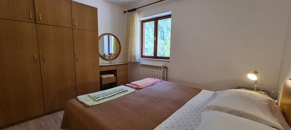 A bedroom with a bed, a nightstand with a lamp, a dresser, a window with curtains, and a radiator. - Apartment Nila (Vrboska)