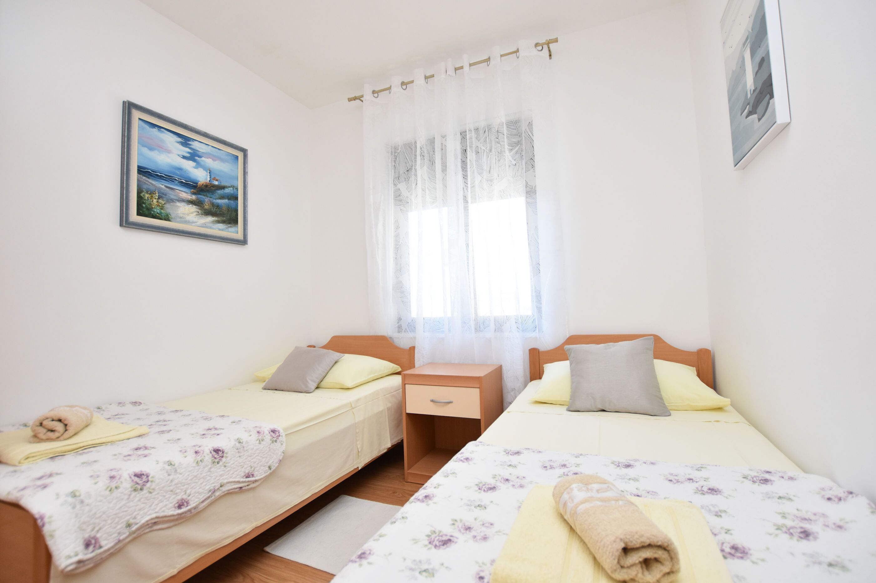 Un dormitorio con dos camas individuales, una mesita de noche, una ventana con cortinas translúcidas y un cuadro en la pared.