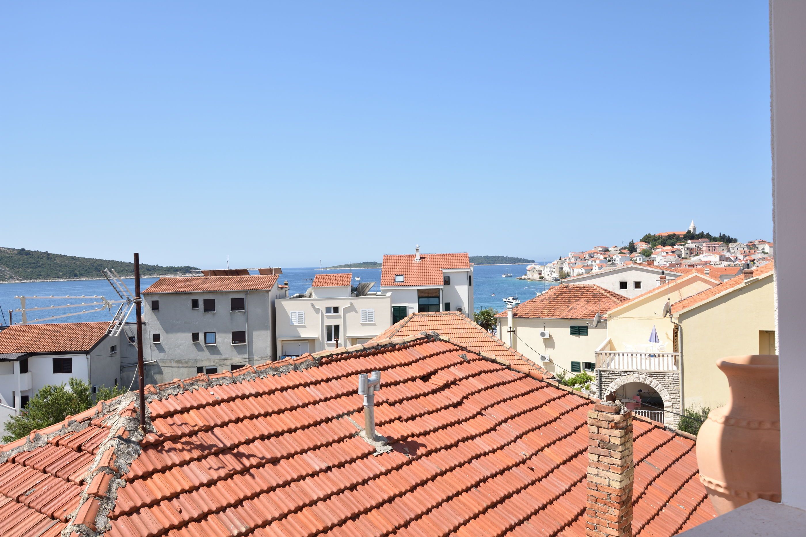 A coastal town with terracotta-roofed buildings, a clear blue sky, and a distant view of the sea.