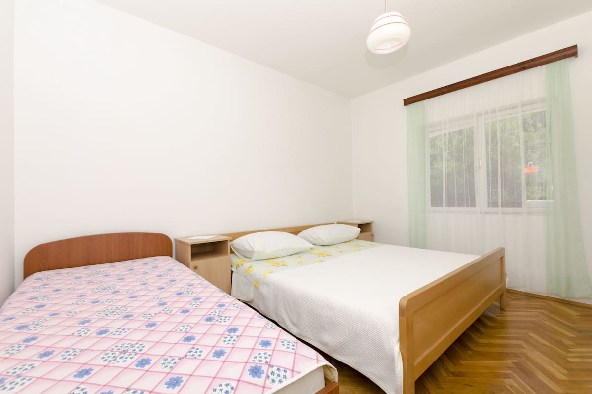 Une chambre avec deux lits, des têtes de lit en bois, une table de chevet et une commode, ainsi qu’une fenêtre ornée de rideaux légers.