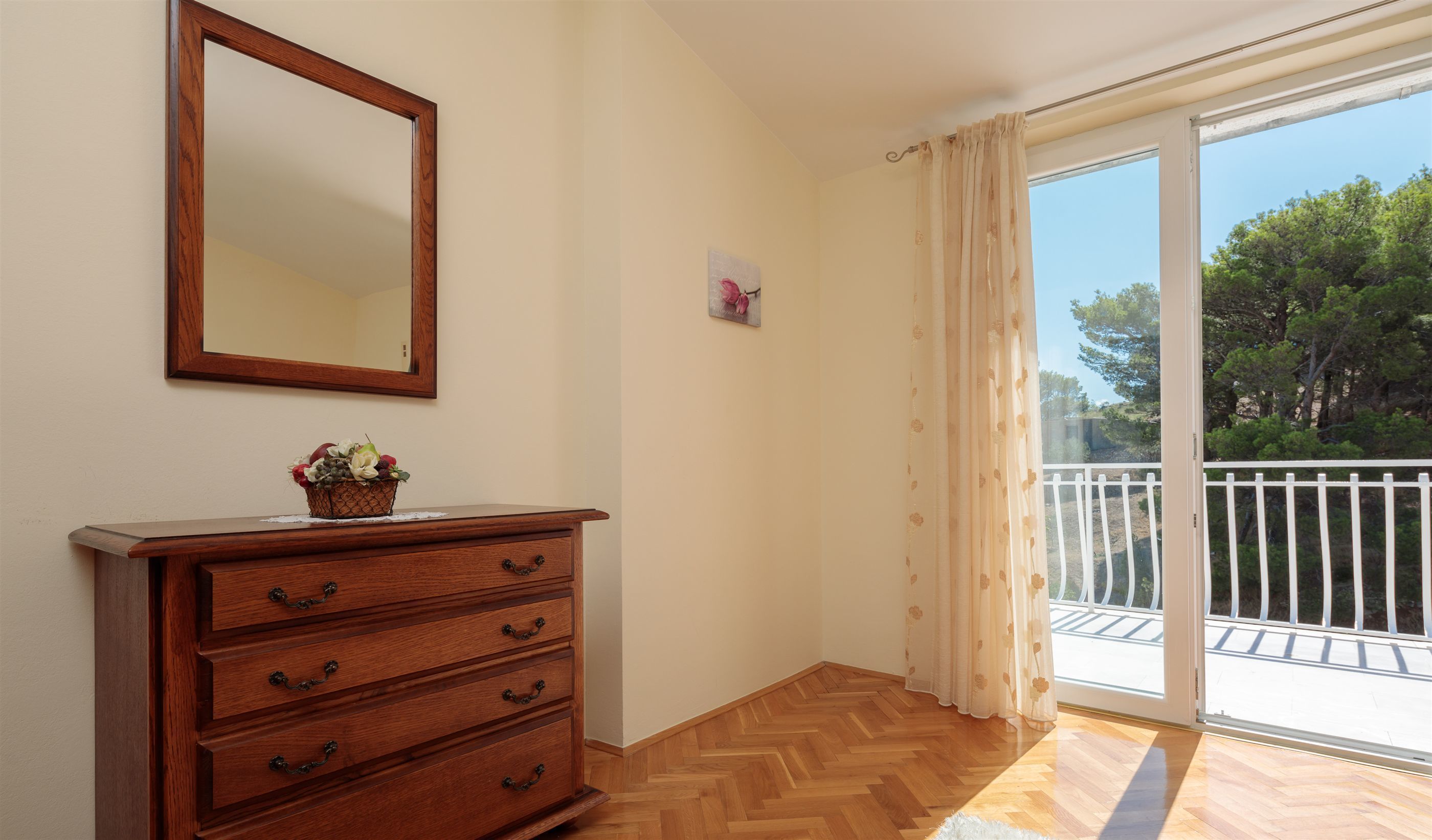 A room with a wooden dresser, a mirror, a small wall decoration, and a balcony with a white railing and trees in the background.
