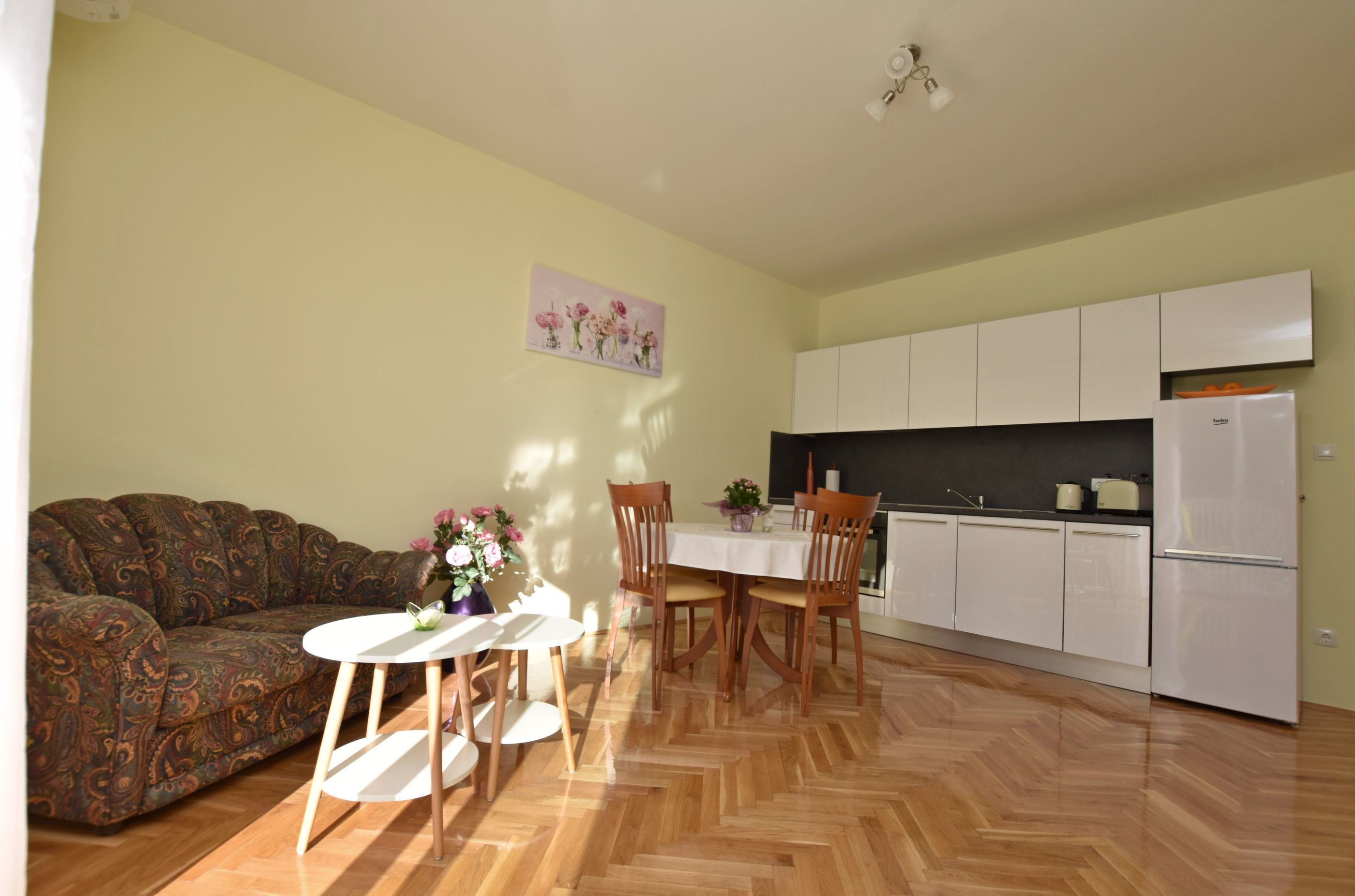 Una sala de estar con sofá, una mesa de comedor con sillas, una cocina con armarios blancos y un refrigerador.